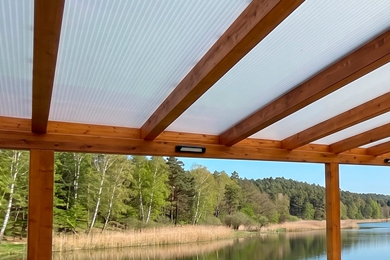 Widok zadaszenia z płyt poliwęglanowych wspartych na drewnianych belkach nad jeziorem i lasem.